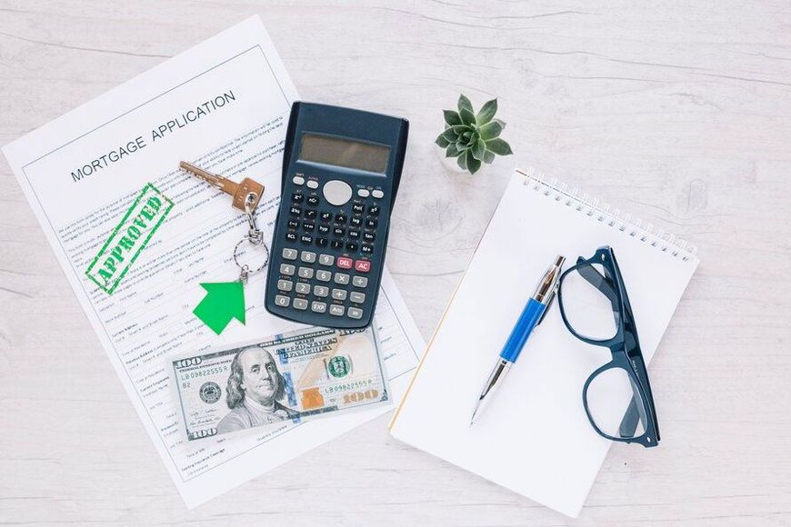 Mortgage application with approval stamp, keys, cash, calculator, notepad, pen, glasses, and a small plant on a white wood table.
