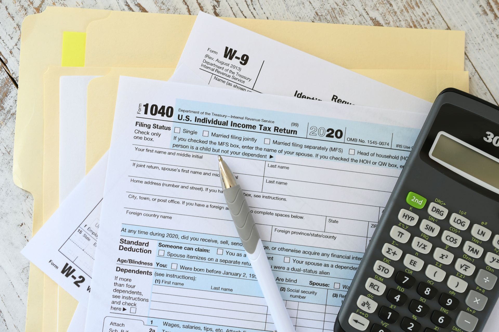 Tax forms and calculator on a wooden table; a pen rests on the largest form.
