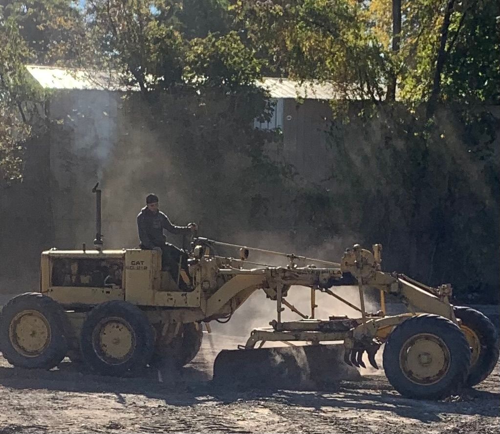 Man operating a yellow road grader, smoothing a dusty gravel surface in an outdoor setting.