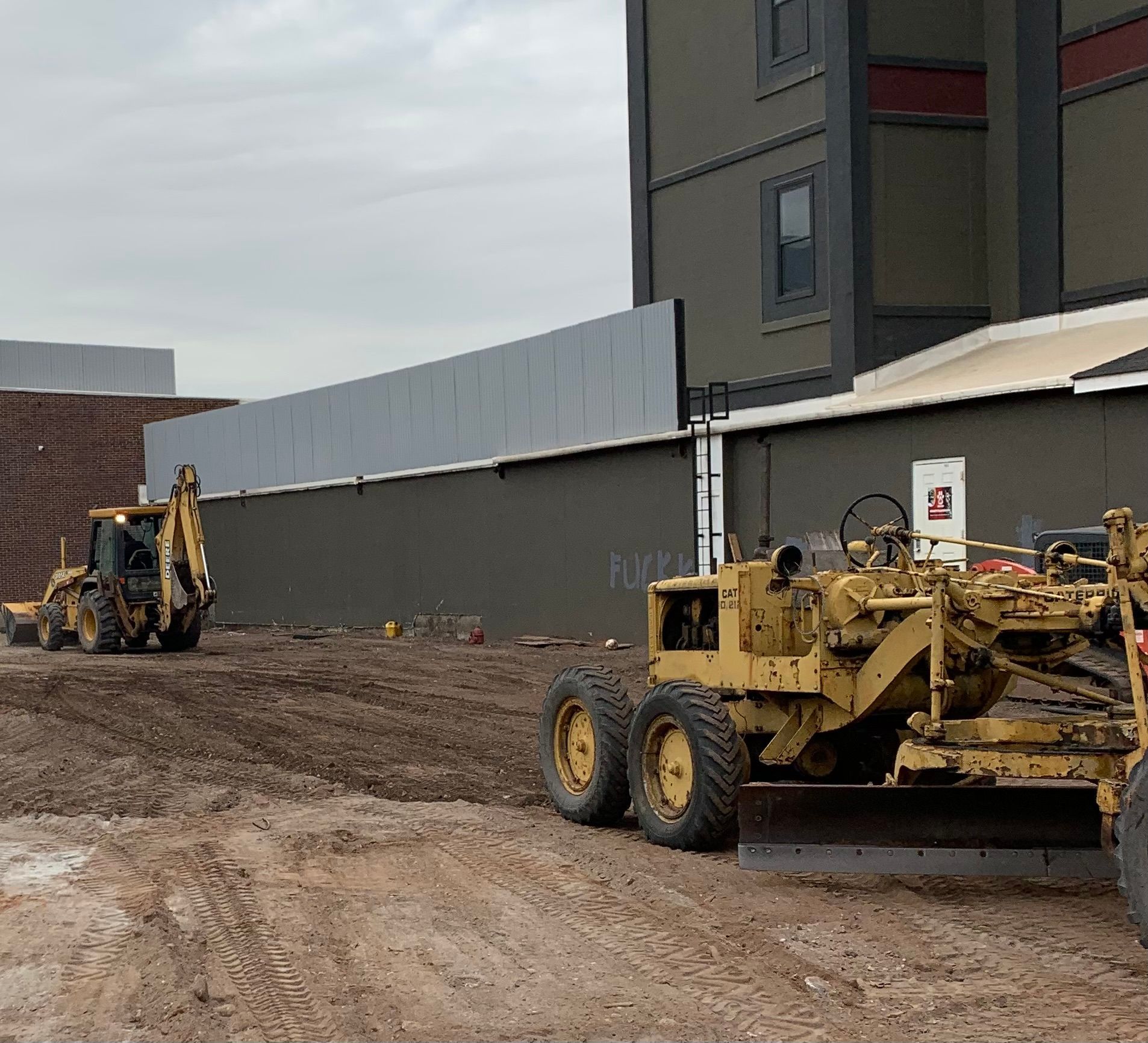 Construction site: yellow grader and backhoe leveling dirt near a building under a cloudy sky.