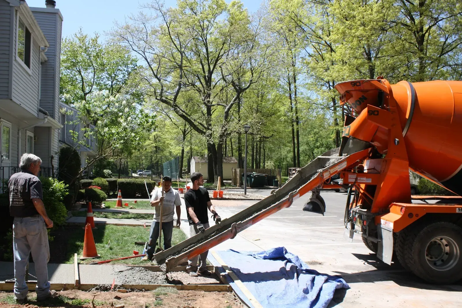 Concrete truck pouring concrete onto a sidewalk in front of a house; three men watch.