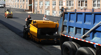 Road paving: Asphalt paver laying fresh asphalt; dump truck alongside, roller in background, sunny day.