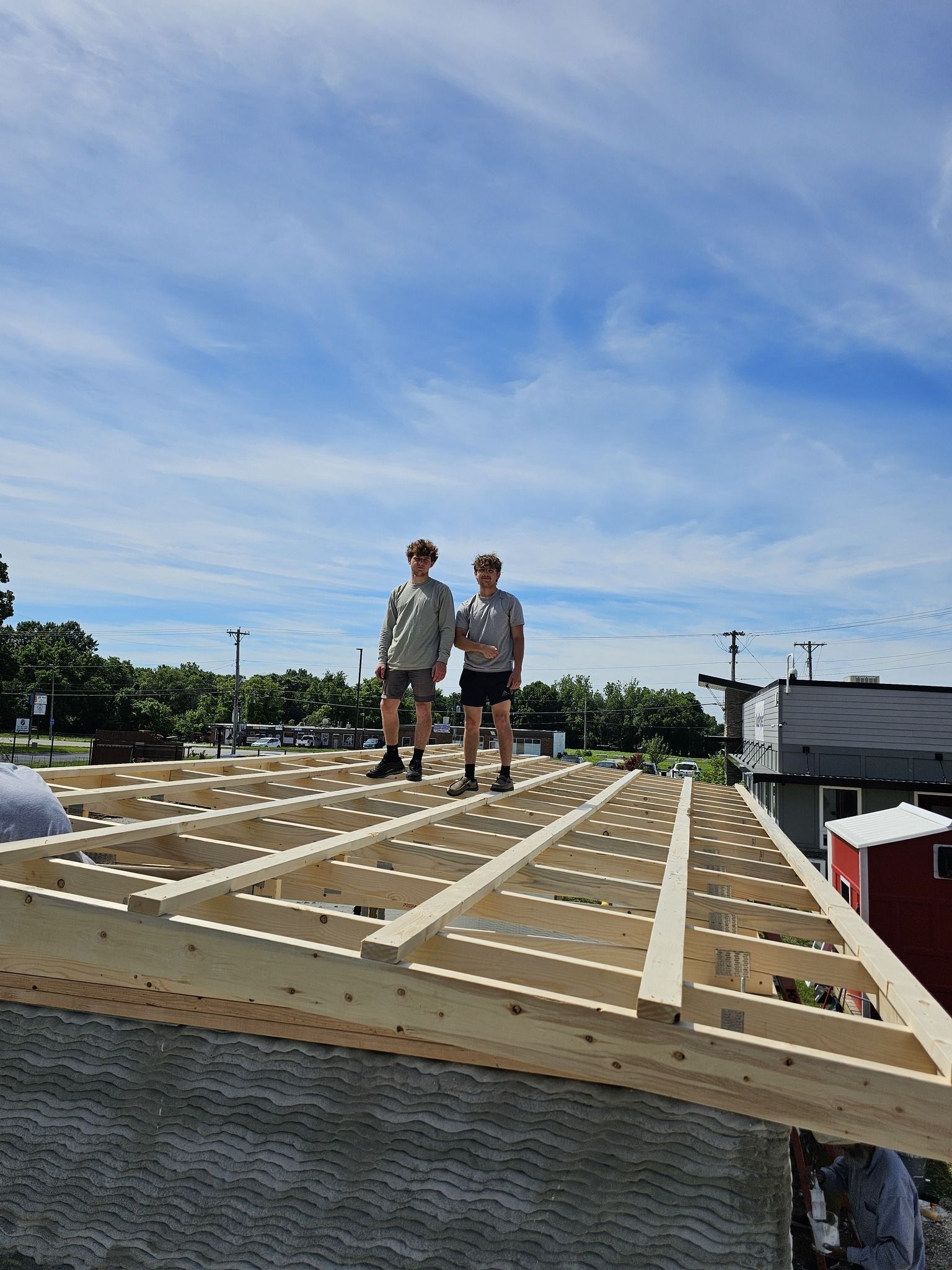 Two people stand on a wooden roof frame under a bright blue, cloudy sky.