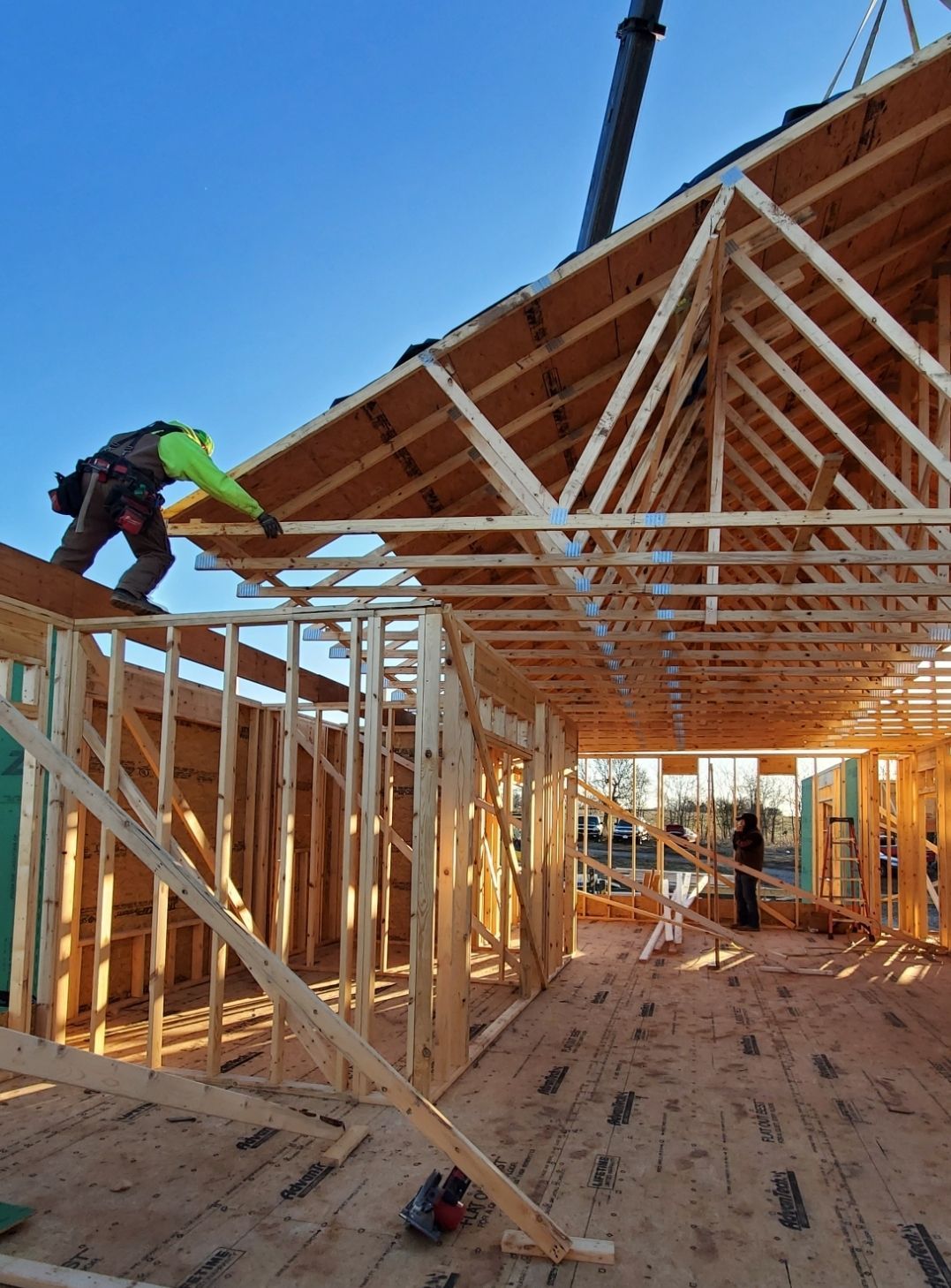 A construction worker in a high-visibility yellow shirt installs wooden roof trusses on a residential building frame.