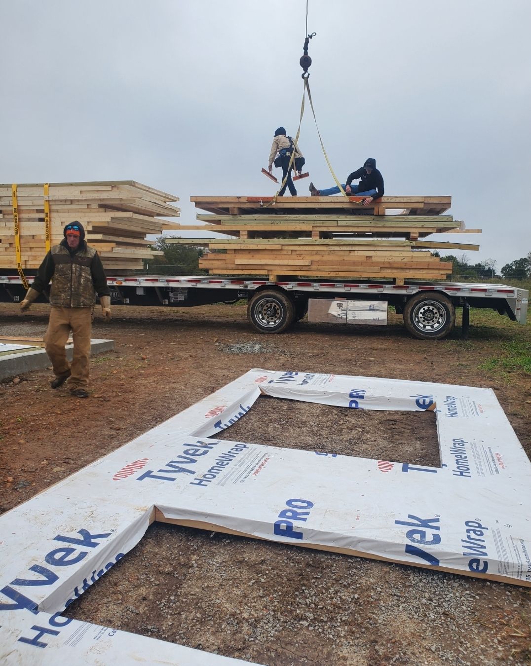 Construction workers on a flatbed trailer being lifted by a crane, with a wall panel resting on the ground nearby.