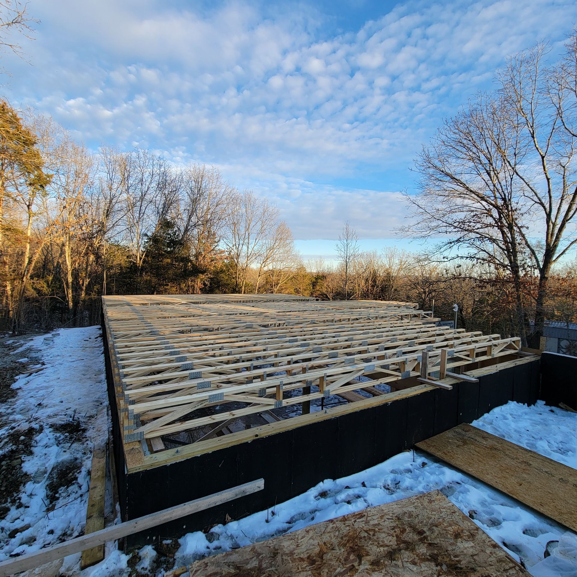 Wooden framing for a house foundation sits atop a black-covered foundation wall in a snowy outdoor landscape.