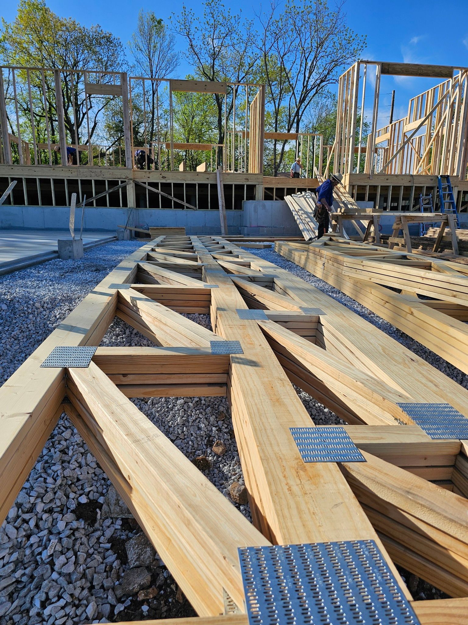 A construction site with wooden roof trusses resting on gravel in the foreground and a partially framed wall behind.