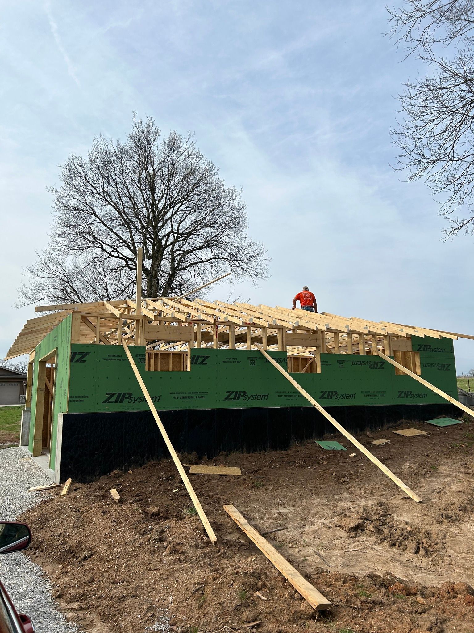 A construction worker stands on the rafters of a building frame covered in green, branded sheathing under a blue sky.