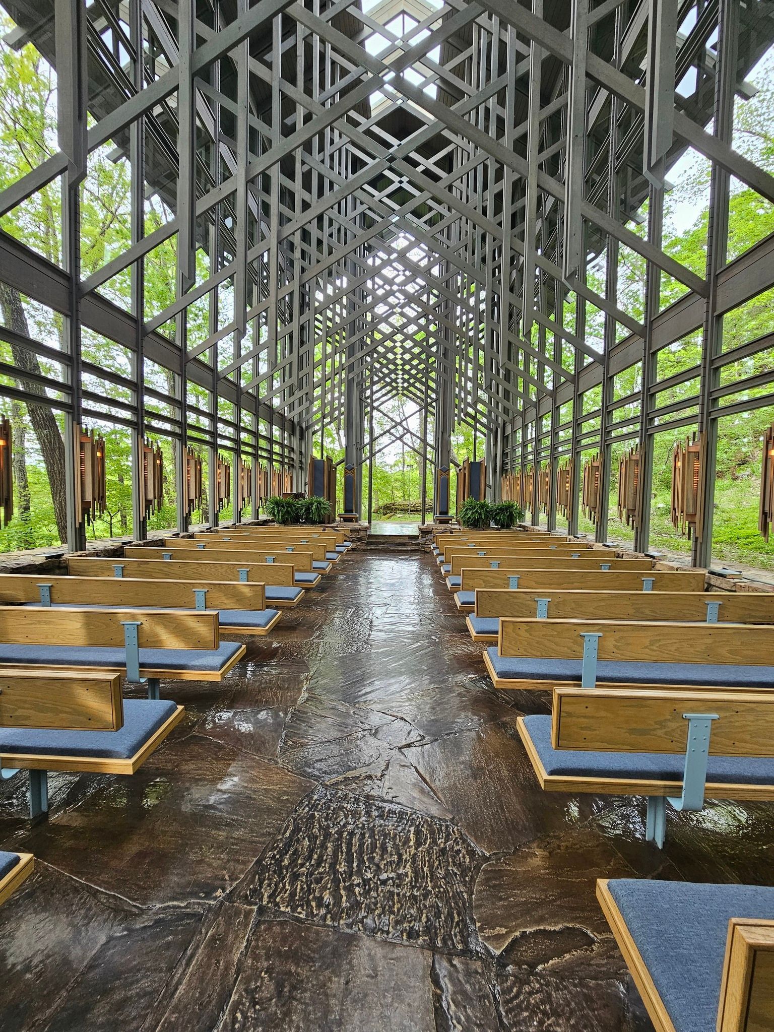 An interior view of a modern chapel featuring a metal truss roof, wooden pews, and a stone aisle, surrounded by greenery.