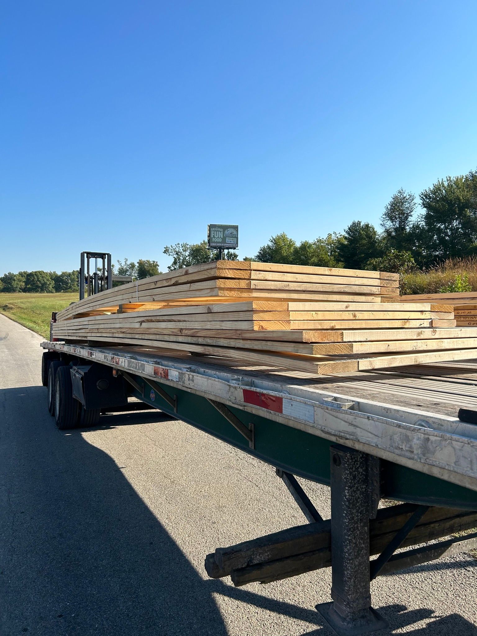 A flatbed trailer loaded with a stack of raw, light-colored wooden planks, parked on a gravel surface under a clear sky.