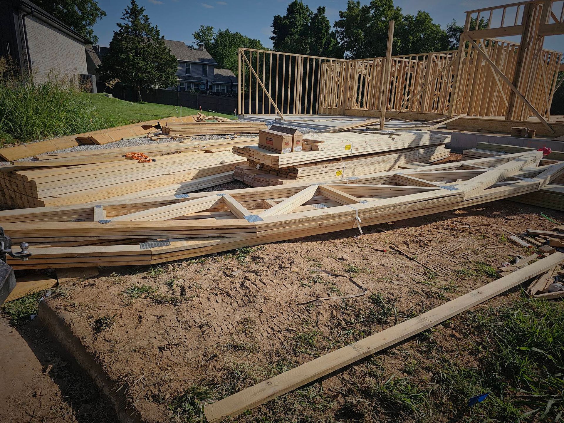 Wooden roof trusses lie on the ground at a residential construction site near a partially framed house structure.