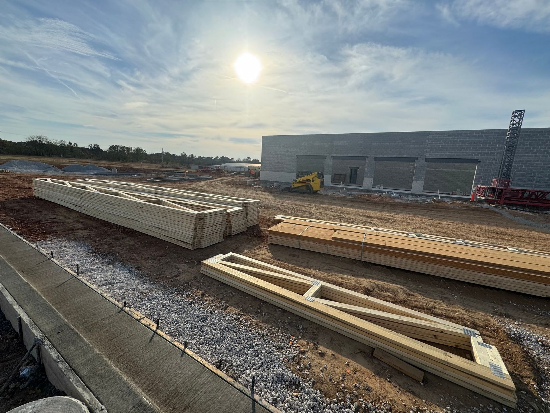 Stacks of wooden roof trusses lie on a dirt construction site near an unfinished concrete building under a bright sun.