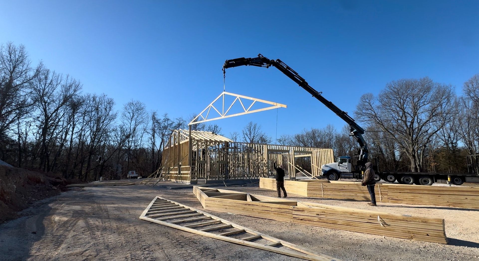 Construction site with a crane lifting a white steel roof frame in a wooded area