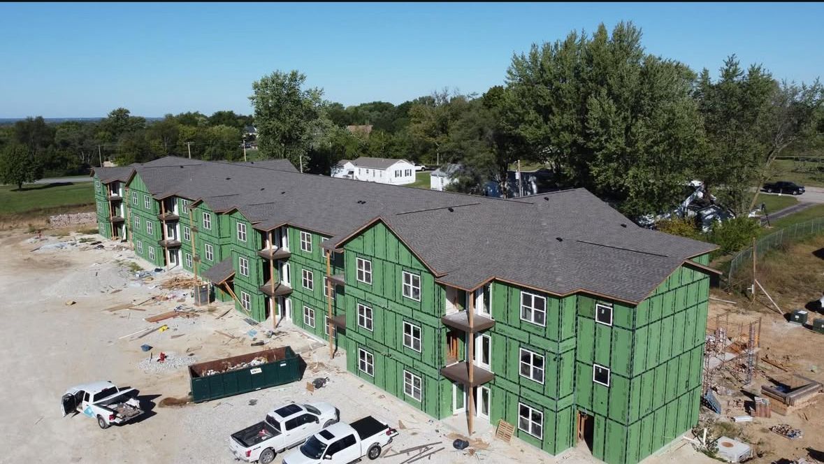 An aerial view of a three-story apartment building under construction, featuring green sheathing and a shingled roof.