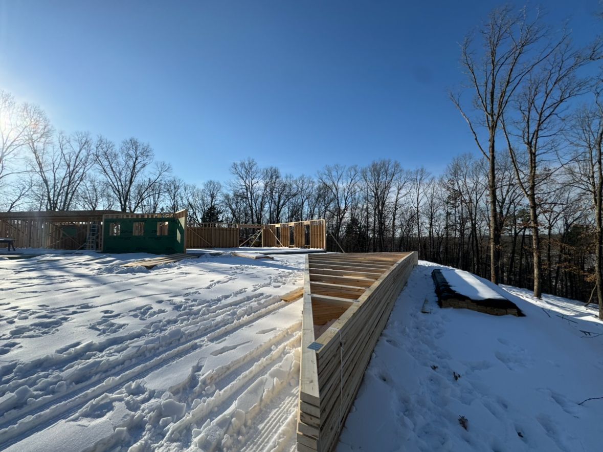 A construction site in the snow, showing a wooden house foundation and a tapered wooden deck edge under a clear blue sky.