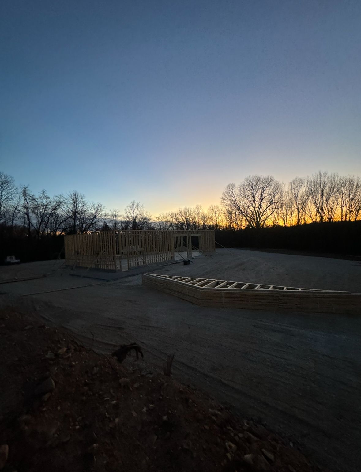Dusk sky over an empty paved lot with a low concrete wall and trees silhouetted on the horizon