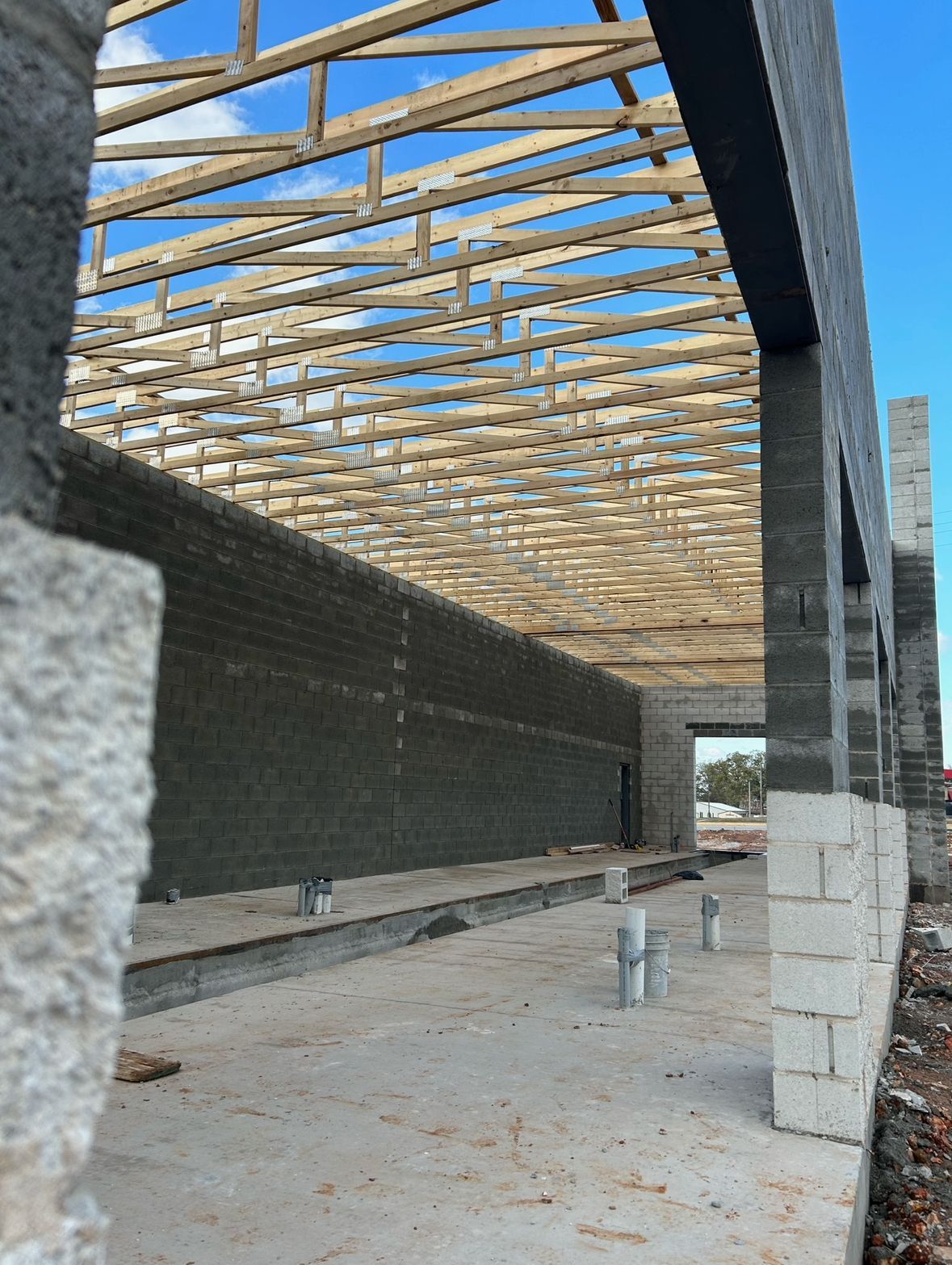 Construction site showing a concrete foundation, dark block walls, and an overhead wooden roof truss structure.