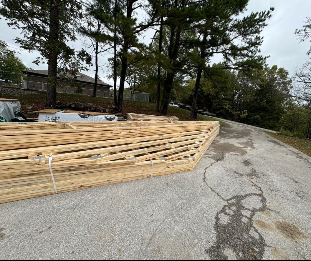 Stacked lumber on a roadside beside a paved drive, with trees and a house in the background.