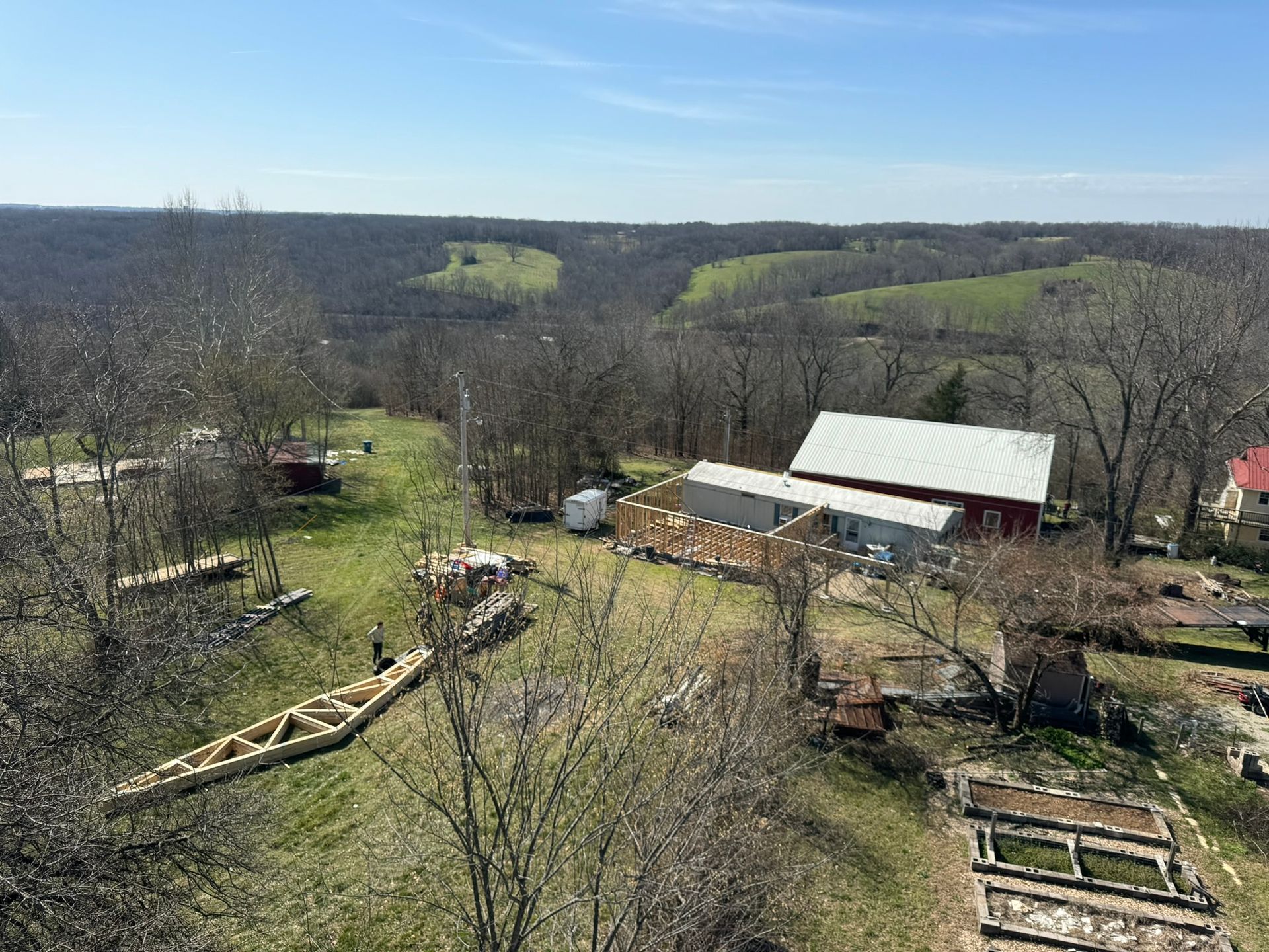 Rural farmyard with barn, sheds, and garden plots on a sunny day overlooking wooded hills