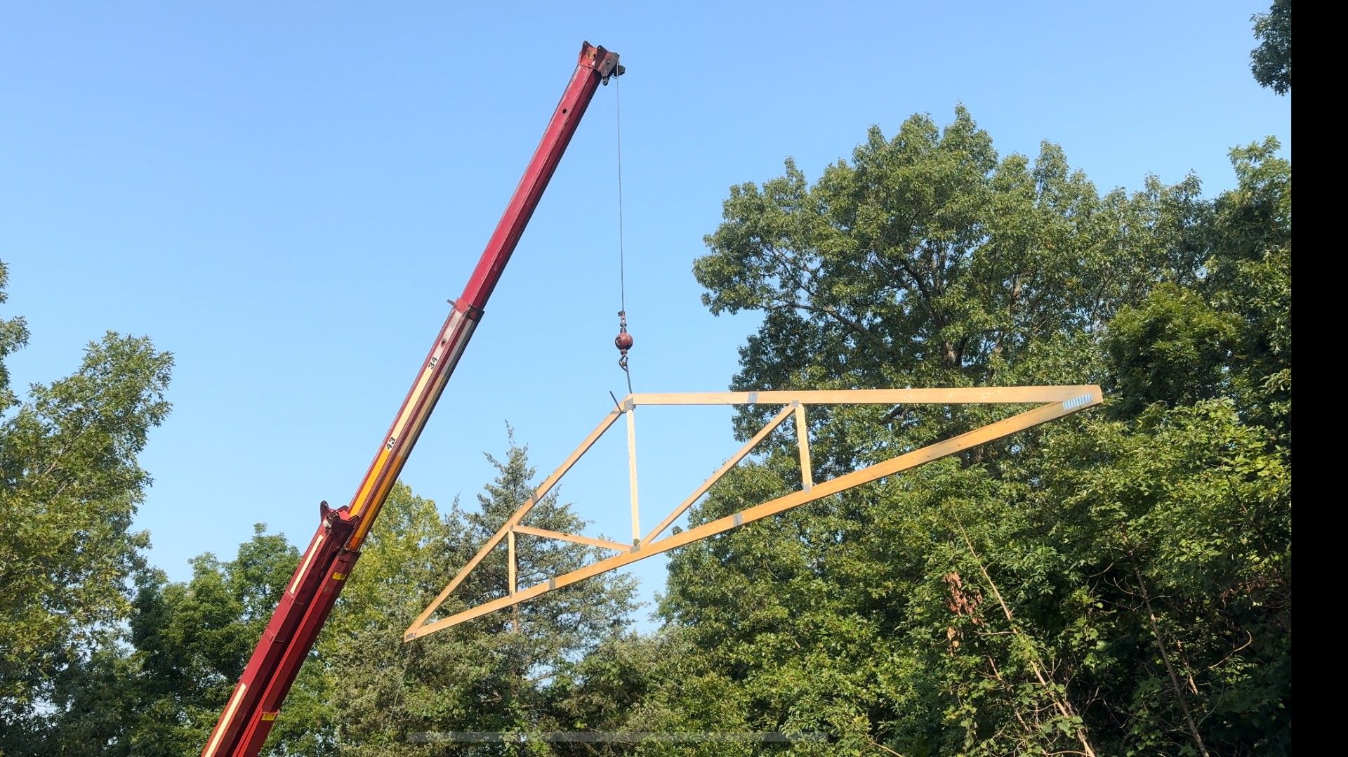 Crane lifting a wooden roof truss above trees on a clear blue day