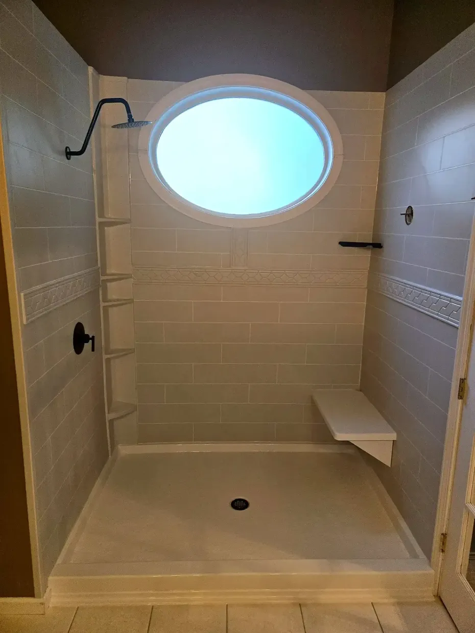 A walk-in shower with light grey tiled walls, a built-in bench, an oval window, and a black showerhead.