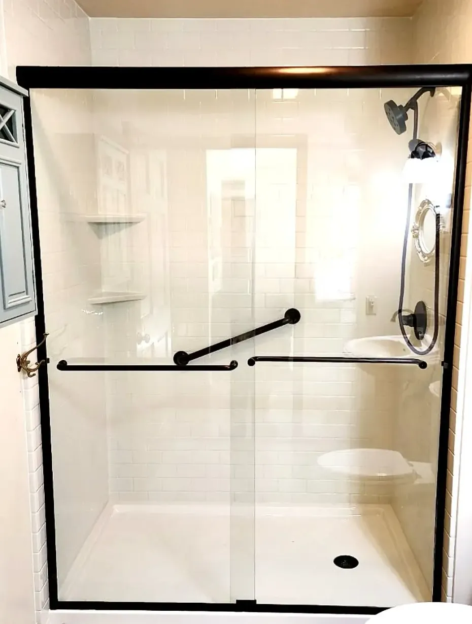 A walk-in shower with clear glass sliding doors, black metal framing, an angled grab bar, and a built-in shower seat.