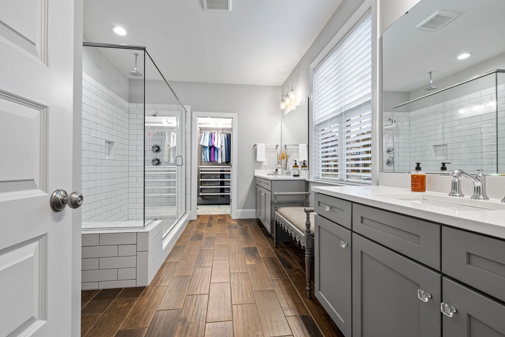 Bright bathroom with white tile, gray vanity, glass shower, wood floor, and walk-in closet in back
