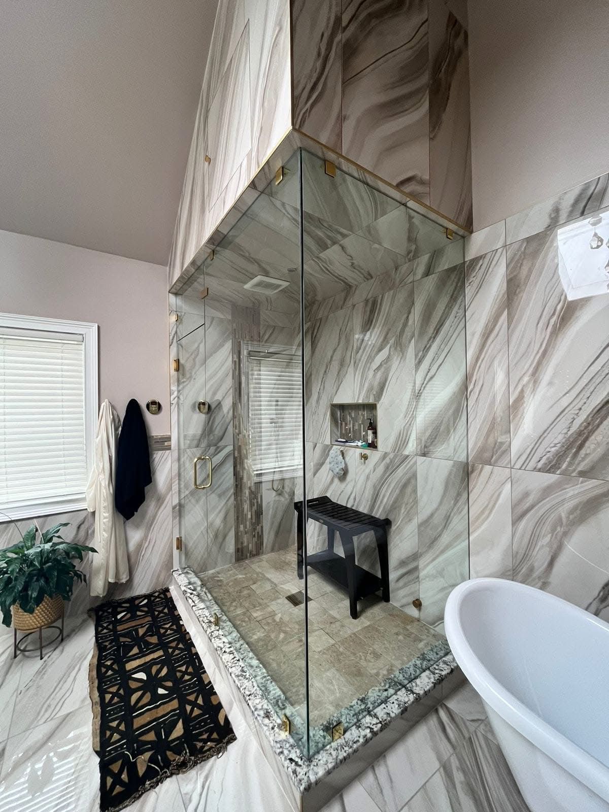 Marble-tiled bathroom with glass shower, white bathtub, black stool, and rug along the wall