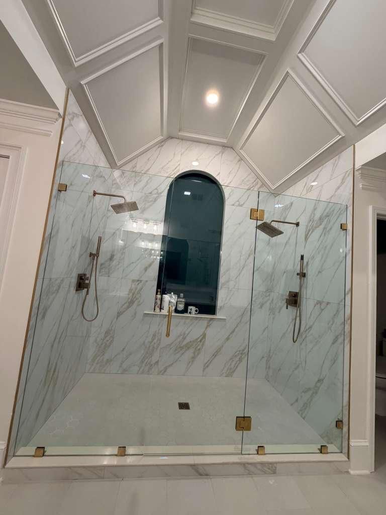 Marble-tiled bathroom with a glass shower enclosure, arched window, and geometric ceiling trim.