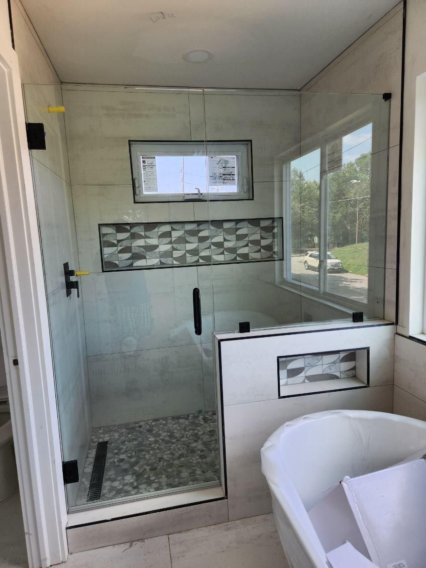 Small tiled shower with glass door, mosaic accent strip, and a white tub in the foreground.