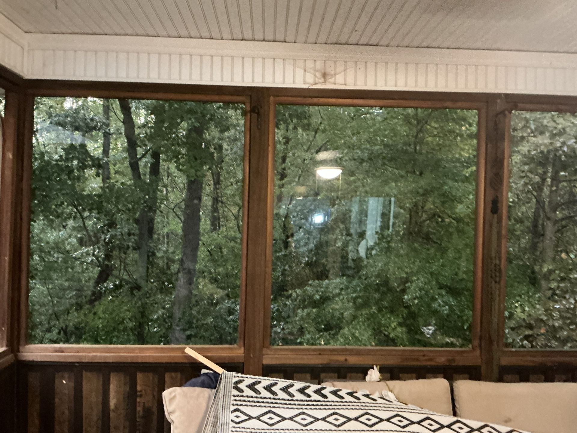 Wooden porch with large windows overlooking dense green forest, with a bench and patterned cushion in the foreground