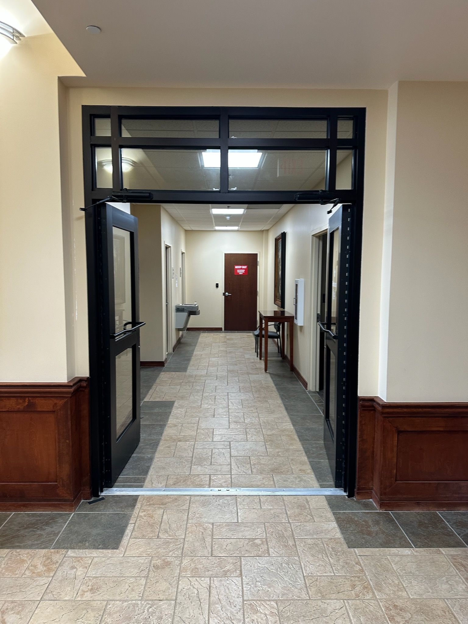 Hallway with tiled floor, wood-paneled walls, and black-framed glass doors leading to a distant red door