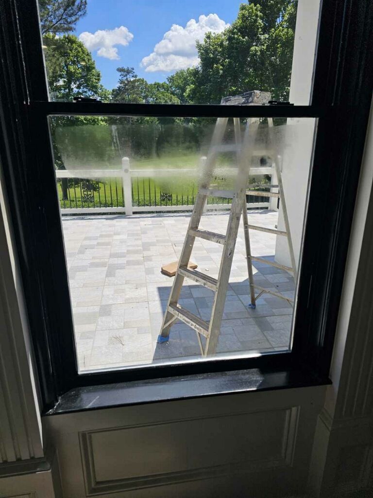 View through a window of a backyard with a ladder, patio, trees, and a fountain sprinkling water.