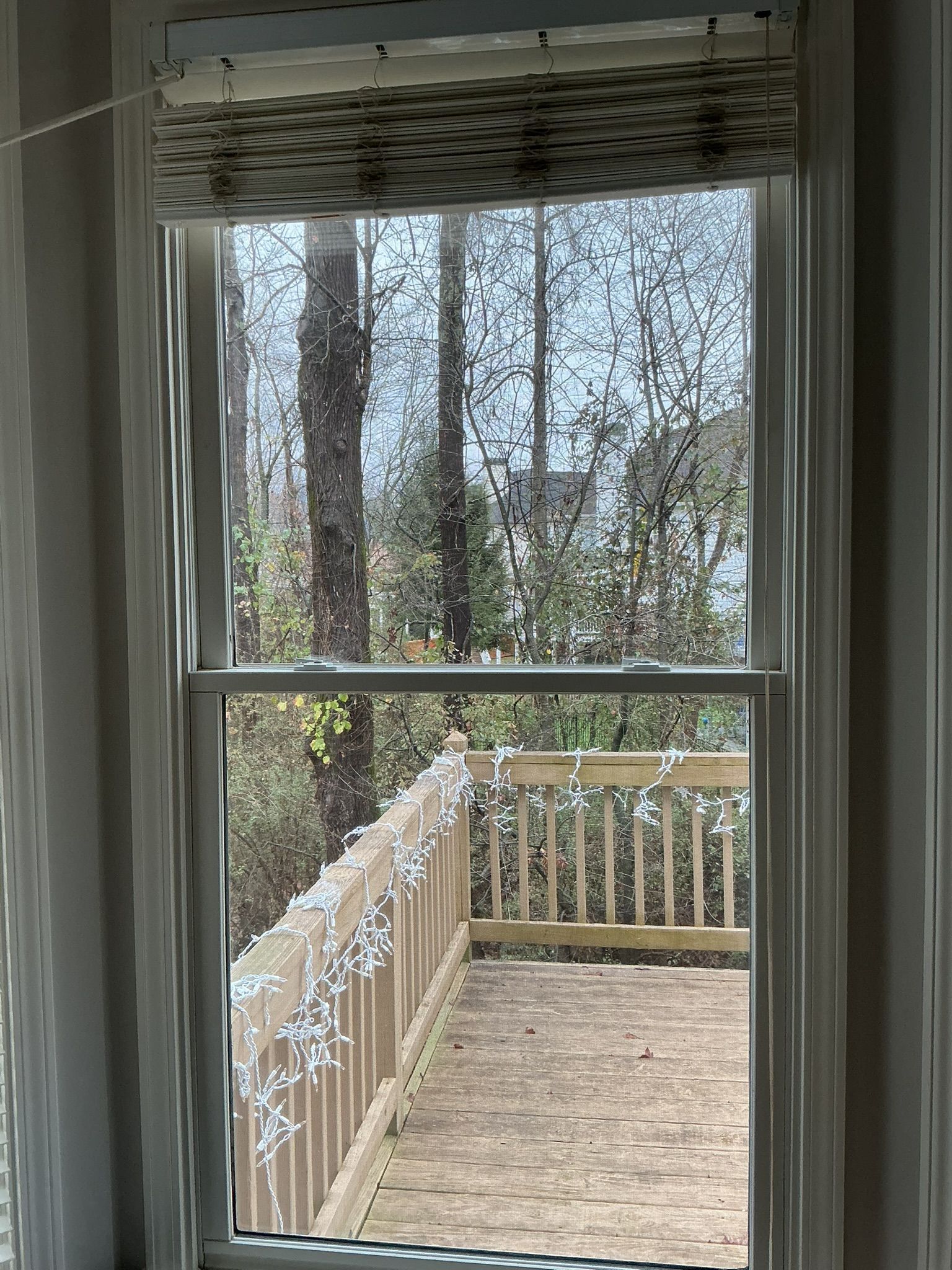 Window view of a wooden deck and railing with string lights, overlooking trees and a lake