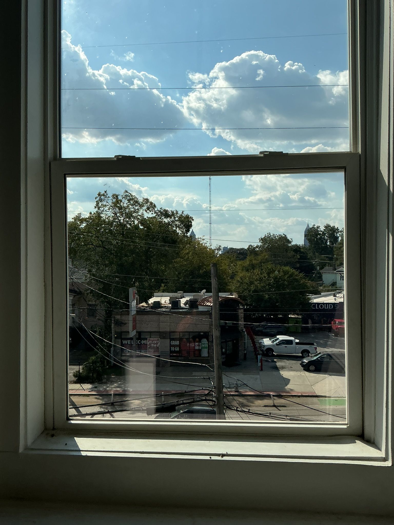 View through a window of a sunny street with trees, parked cars, and a partly cloudy sky