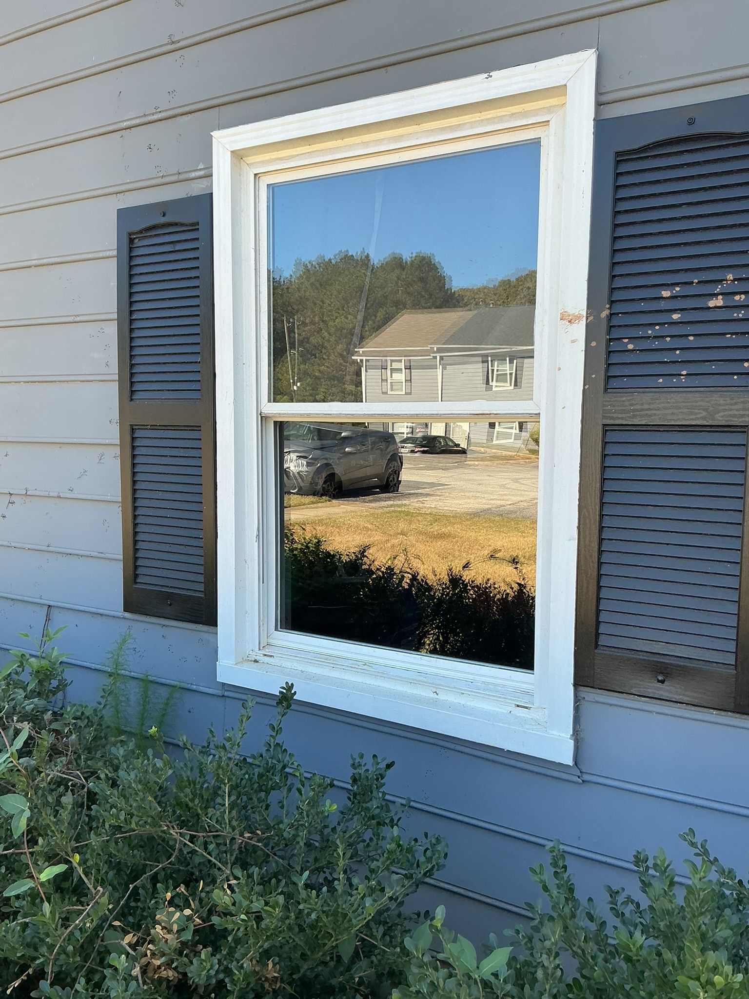 White-trimmed window on blue house siding reflecting a yard and neighboring home, with shutters on both sides