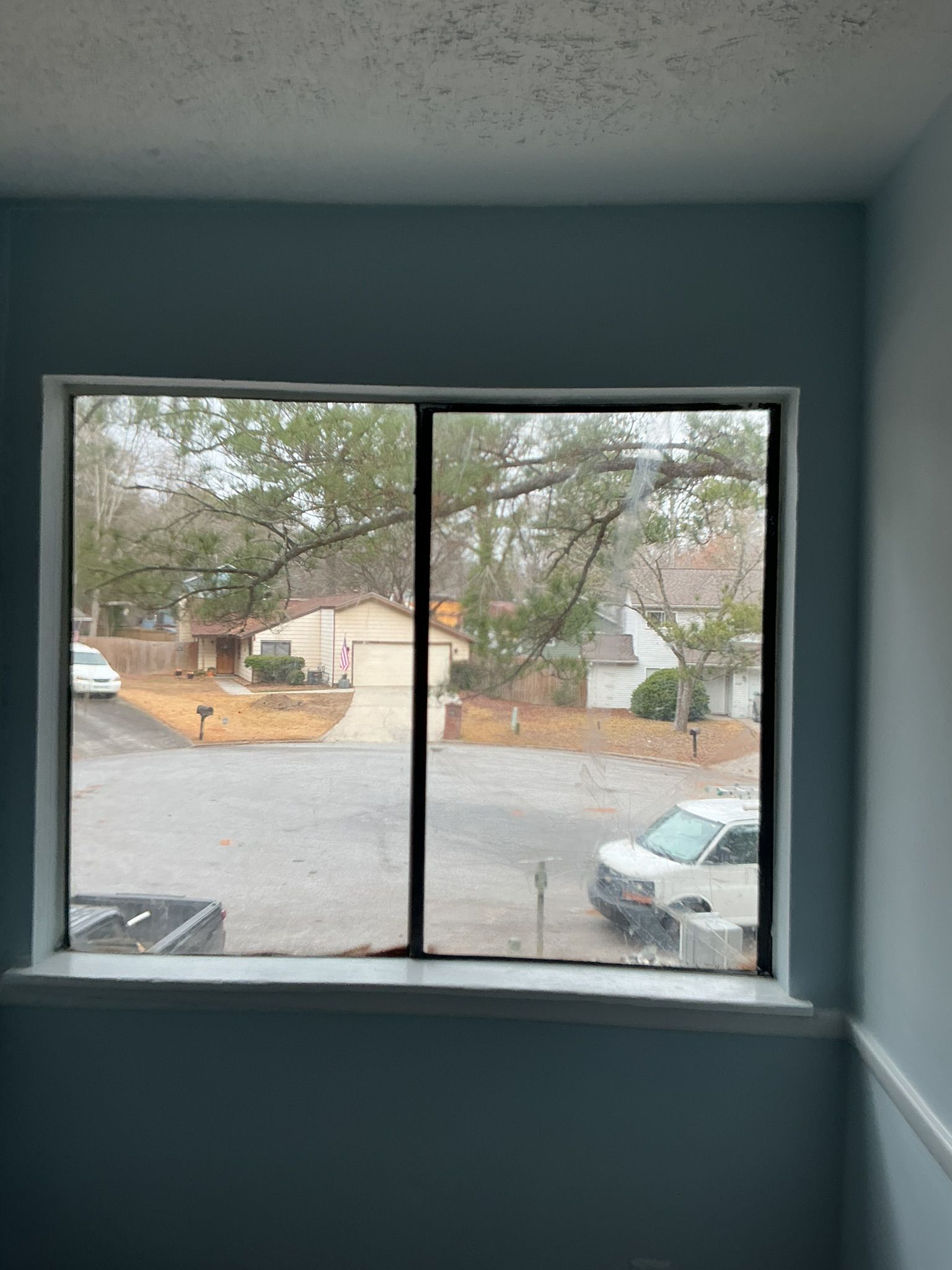 View through a window to a residential street with parked cars and houses, framed by blue-gray interior walls.