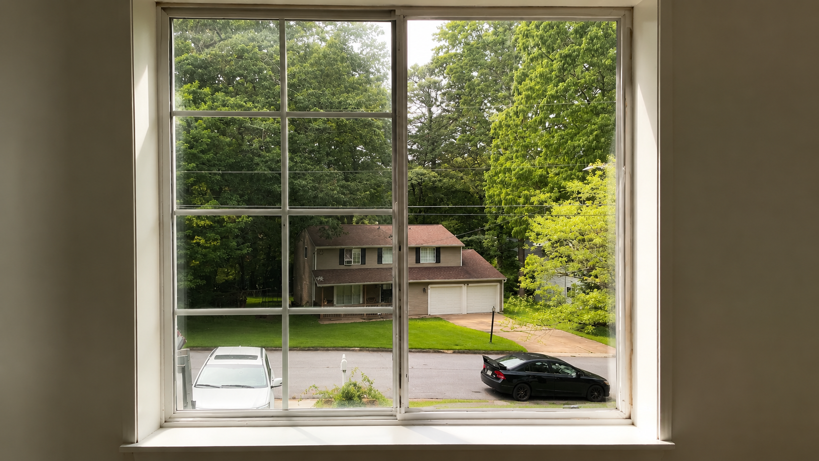 View through a window of a suburban house and street, with two parked cars and trees outside.