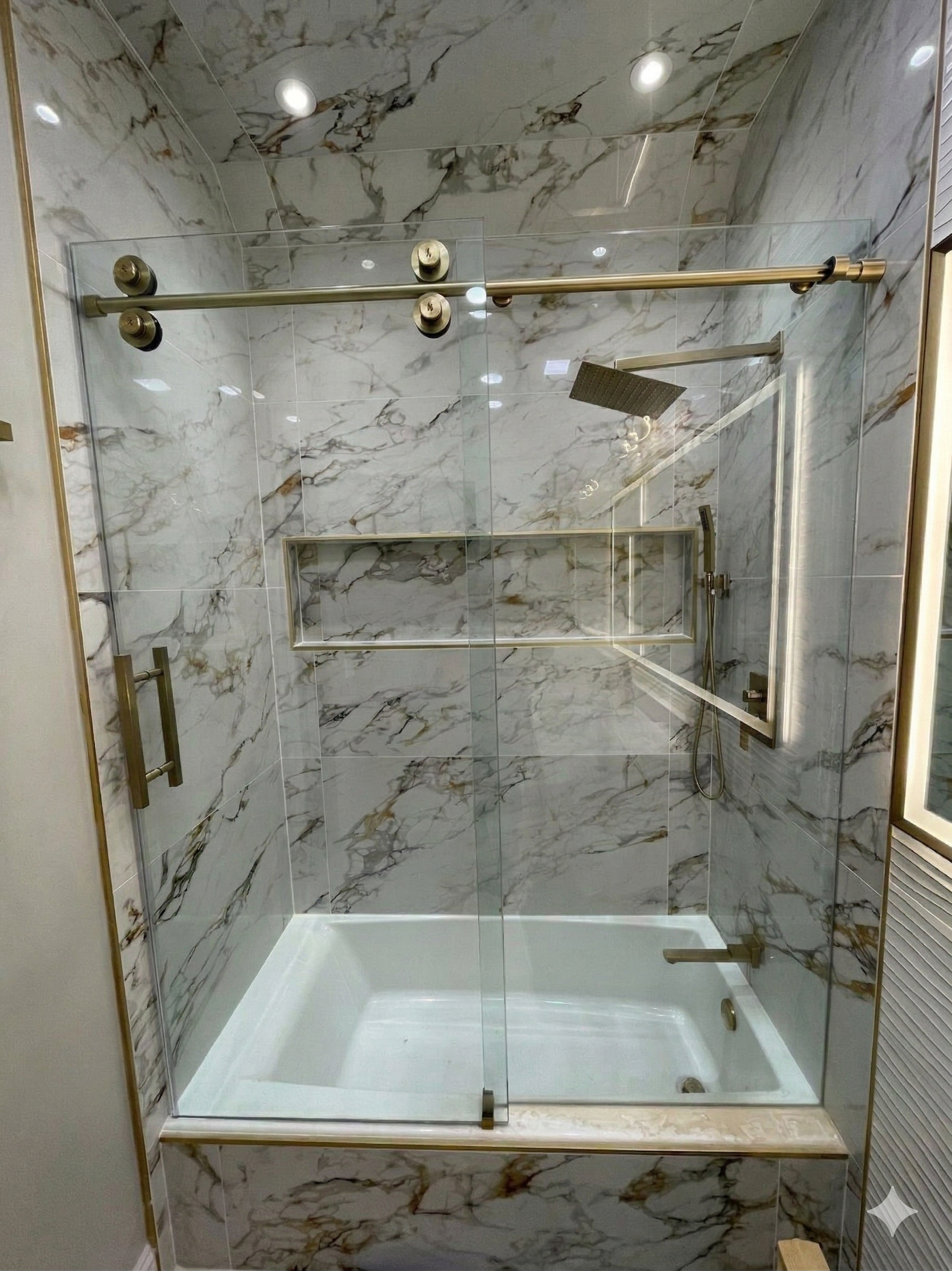 Marble-tiled shower stall with glass door, brass hardware, built-in shelf, and white bathtub