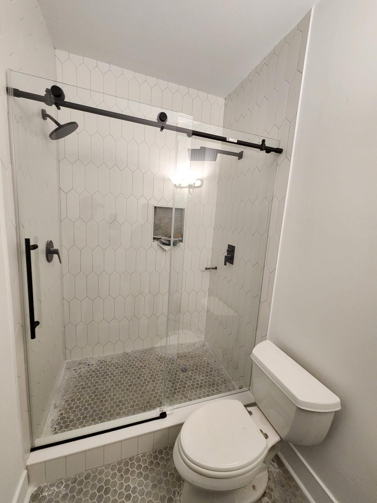 Small tiled bathroom with glass walk-in shower, black hardware, wall mirror, sink, toilet, and mosaic floor.
