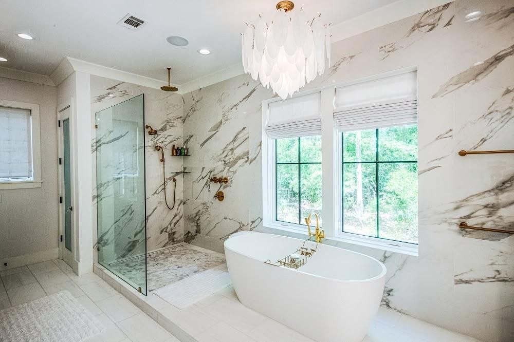 Bright luxury bathroom with glass shower, freestanding tub, marble walls, and large window.