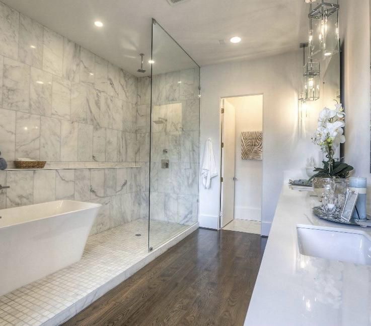Modern bathroom with glass shower, white tub, marble walls, and dark wood floor