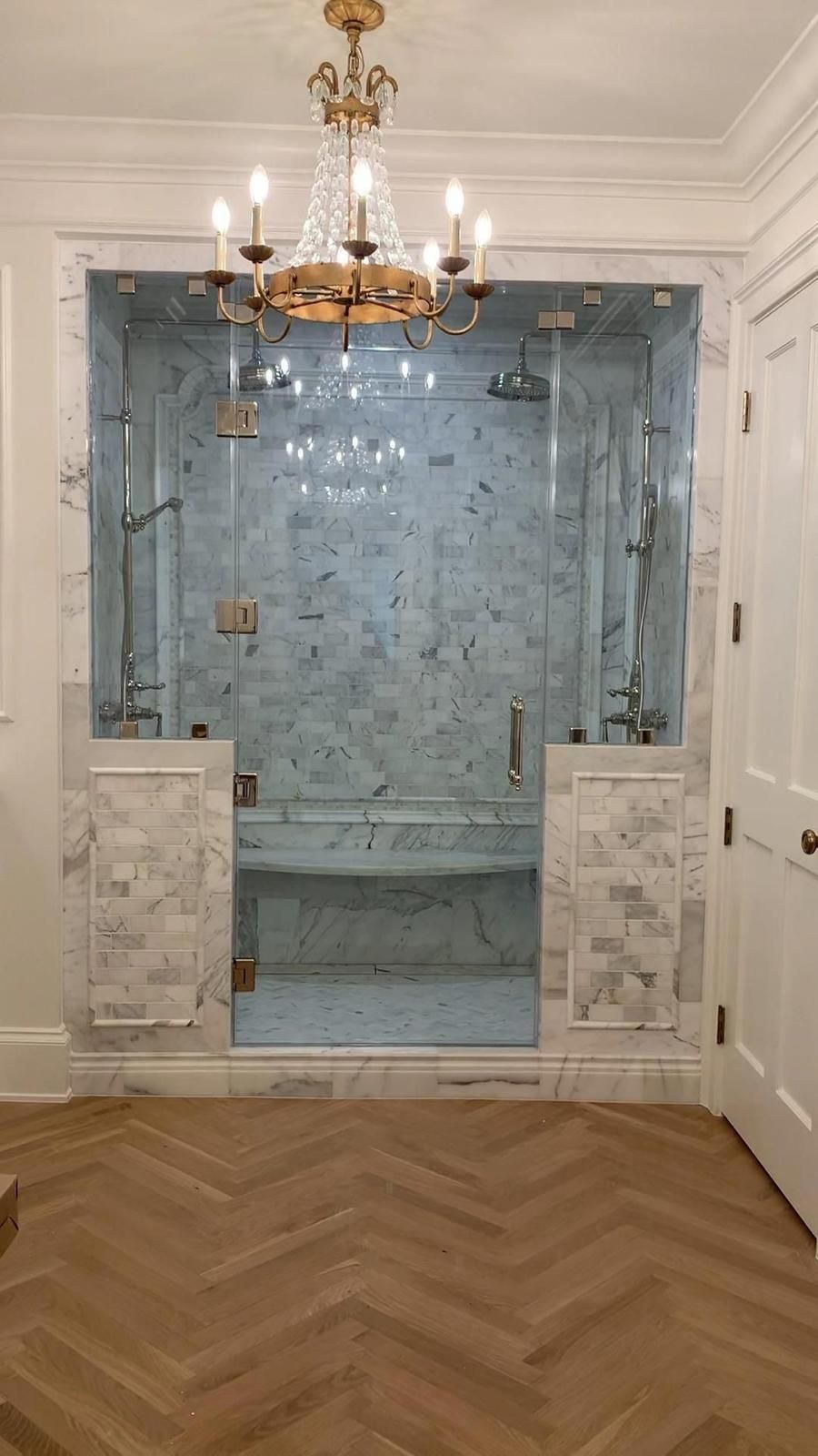 Elegant bathroom with blue-tiled glass shower, marble half walls, chandelier, and herringbone wood floor