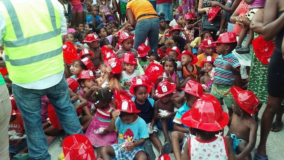 Children in red hard hats crowd around a worker in a safety vest at an outdoor event