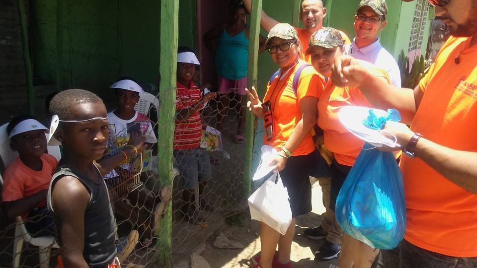 Volunteers in orange vests hand out supplies to people standing by a fence outdoors
