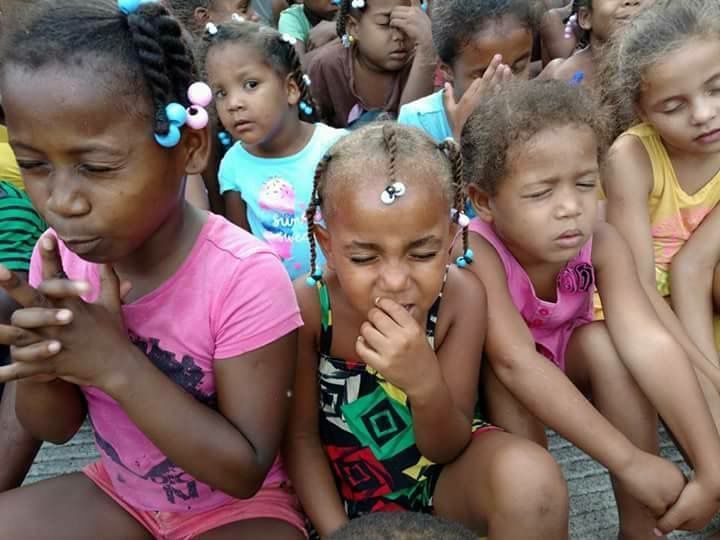 Several children sit closely together, some with eyes closed and hands near their faces, in colorful clothing.