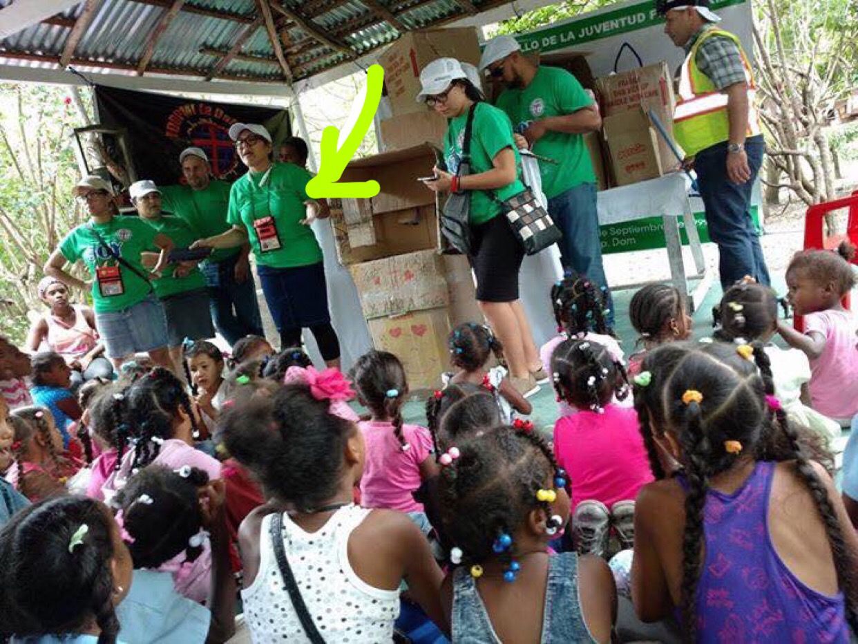 Green-shirted volunteers on a stage handing out items to a crowd of children outdoors