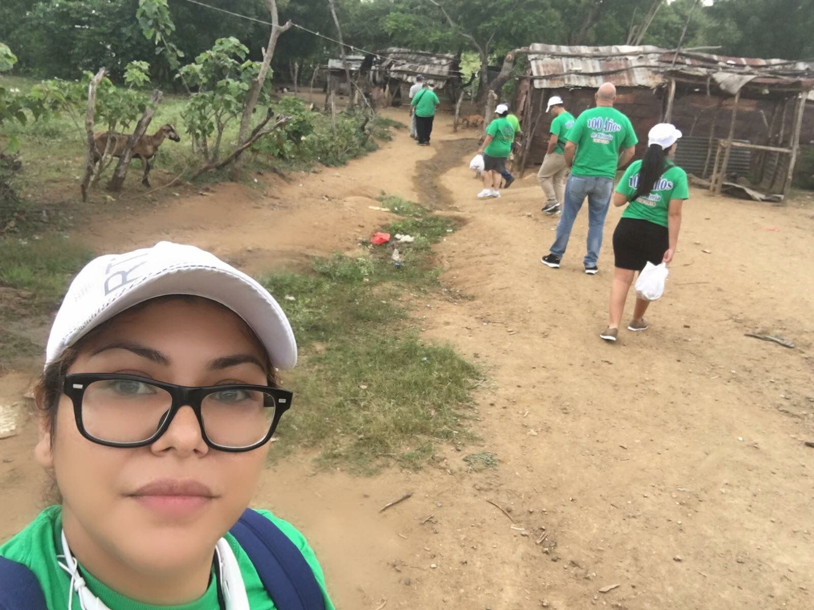 Group in green shirts walking along a dirt path outdoors, with a woman taking a selfie in the foreground.