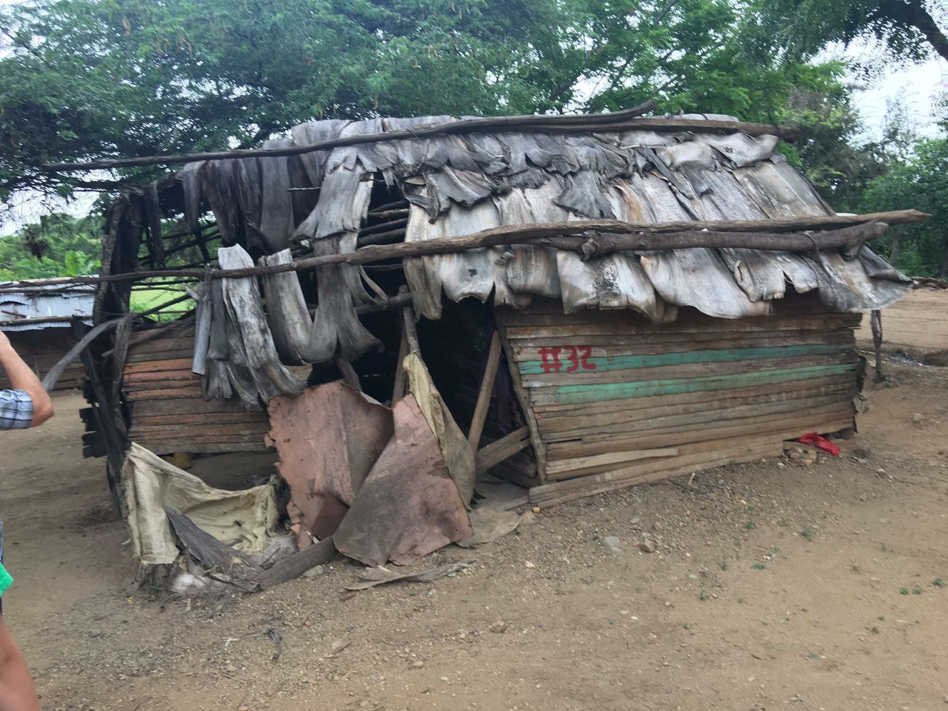 Makeshift hut with torn tarp roof and wooden walls on a dirt clearing, surrounded by trees.