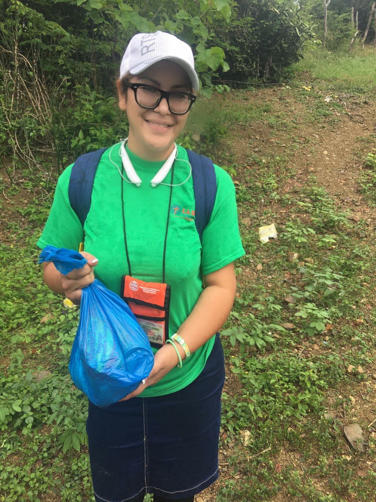 Person in a green shirt and white cap holding a blue bag on a muddy trail outdoors